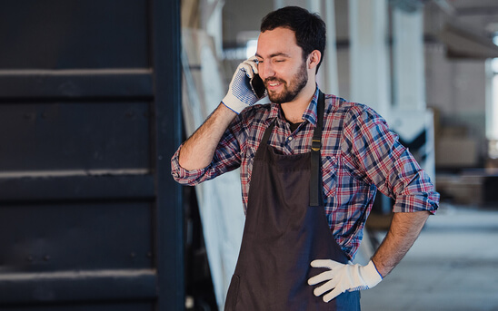 young gentleman wearing a brown apron and garden gloves talking to someone on his mobile phone